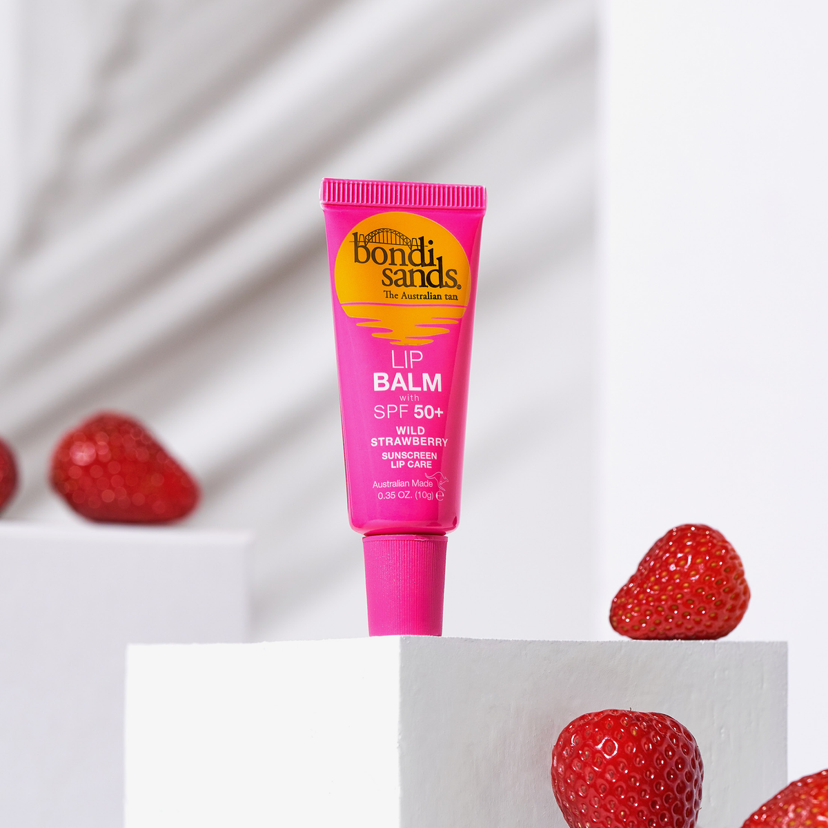 Eine rosafarbene Tube Lippenbalsam LSF 50+ mit Erdbeeren liegt auf weißen Blöcken vor einem grau-weißen Hintergrund.