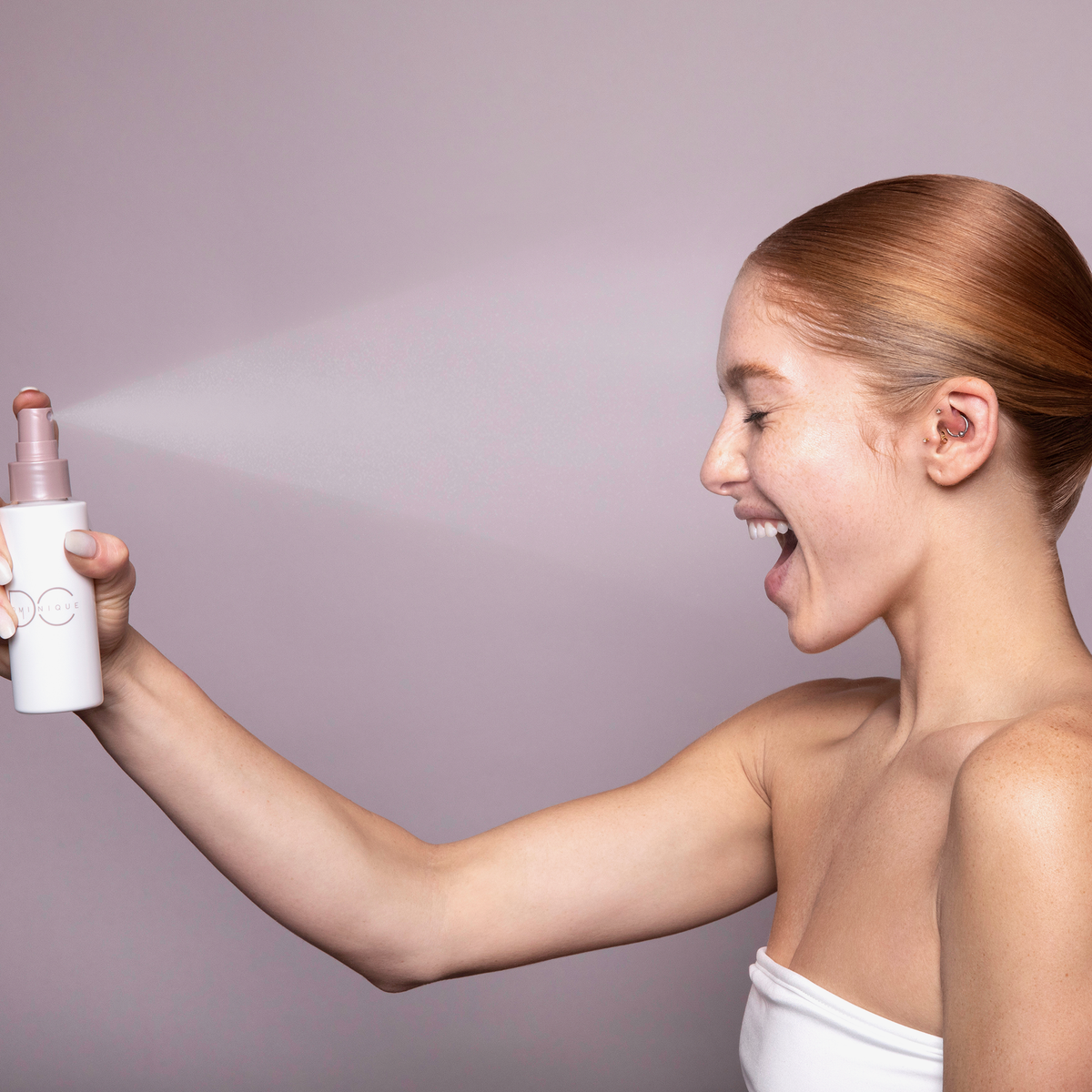 Eine lächelnde Frau sprüht sich vor einem hellrosa Hintergrund aus einer Flasche Ultra Hydrating Fine Mist ins Gesicht.