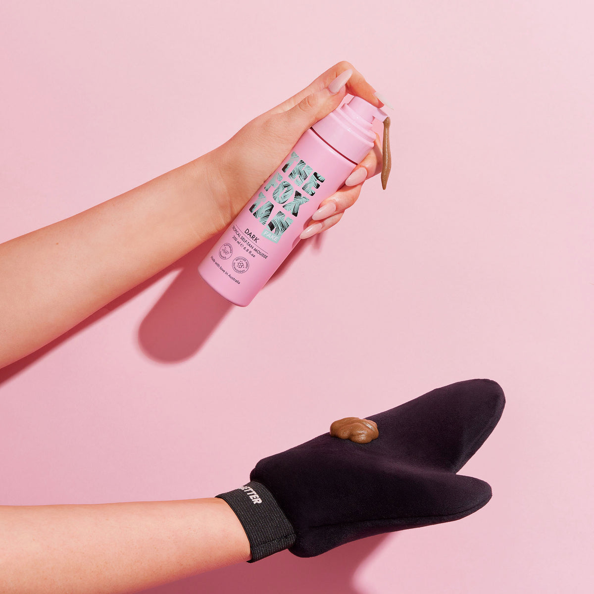 Hände, die das Dark Tropical Self-Tan Mousse aus einer rosa Flasche auf einen schwarzen Handschuh vor einem rosa Hintergrund auftragen.