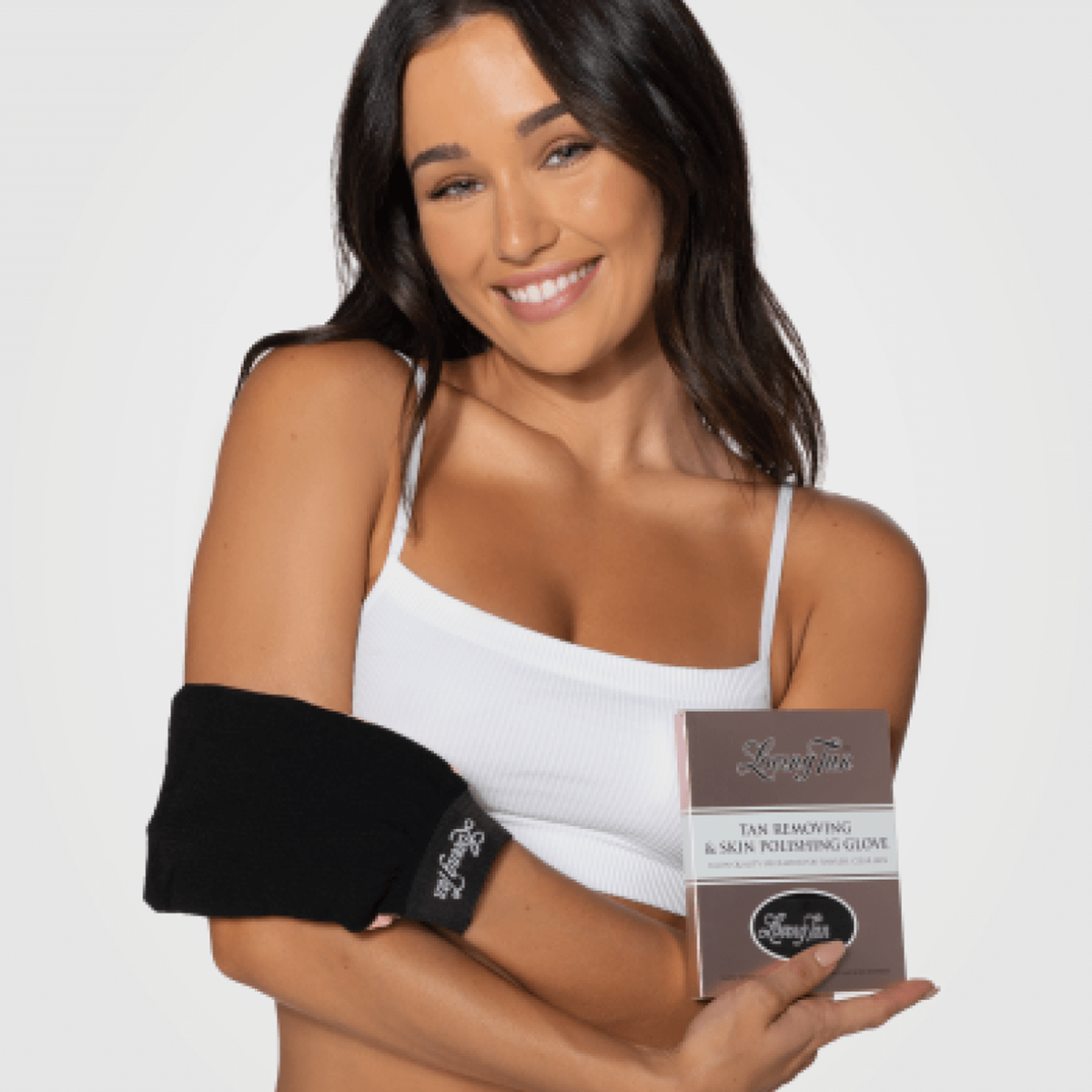 Eine lächelnde Frau in einem weißen Tanktop hält den Tan Removing & Skin Polishing Glove To Remove Self Tan mit Verpackung in der Hand.