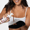 Eine lächelnde Frau in einem weißen Oberteil trägt Loving Tan Bronzing Mousse auf einen schwarzen Handschuh auf.