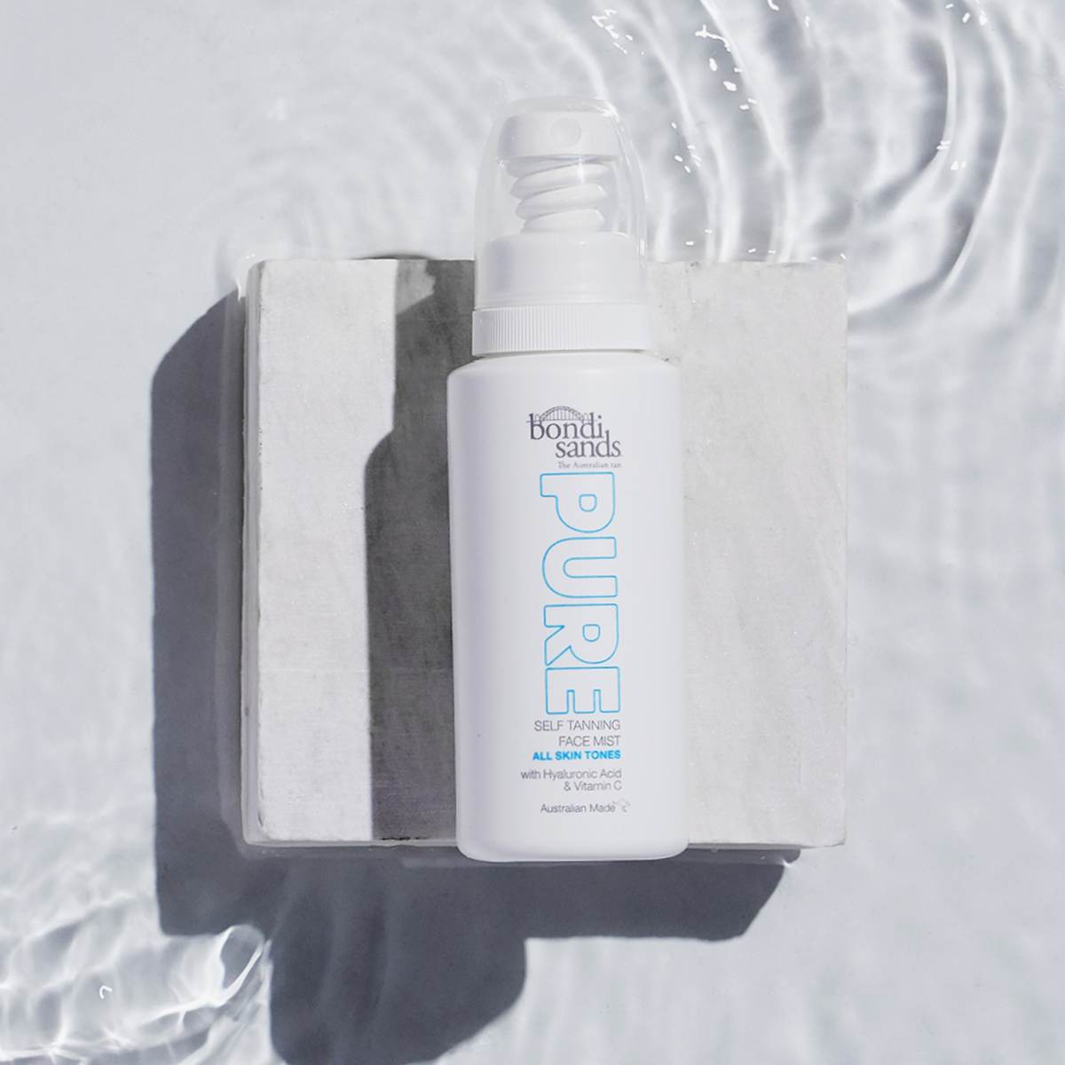 Eine strukturierte Oberfläche mit einer weißen Flasche Pure Tanning Face Mist von Bondi Sands, umgeben von plätscherndem Wasser