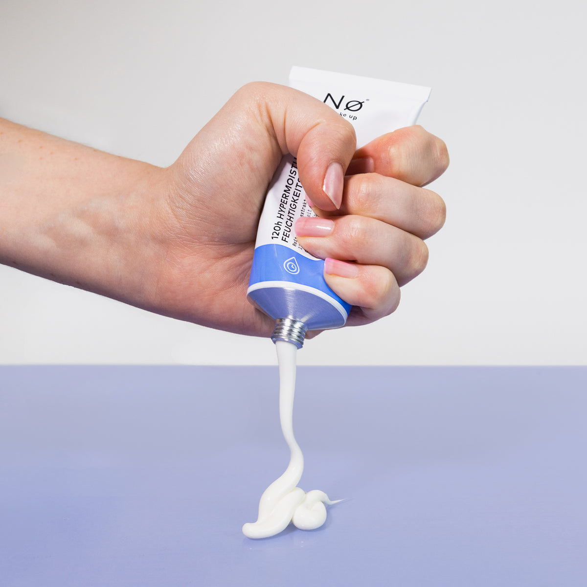 Eine Hand drückt aus der rain tøday 120h mattierende Feuchtigkeitscreme-Tube, eine weiße Creme auf eine hellviolette Oberfläche.