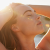 Frau mit geschlossenen Augen genießt die Sonne, hält die Hände im Haar. Goldenes Sonnenlicht beleuchtet ihr Gesicht und erzeugt warme Lichtreflexe.
