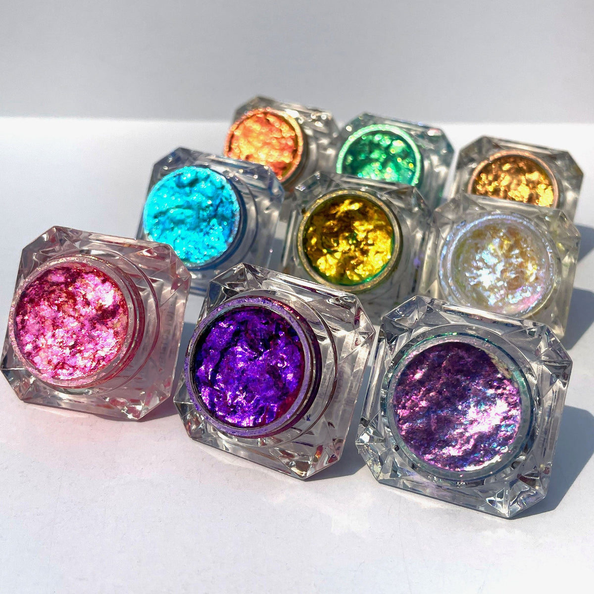 9 bunte Tiegel Infinite Chrome Flakes liegen geöffnet und von der Sonne angestrahlt auf weißer Oberfläche.