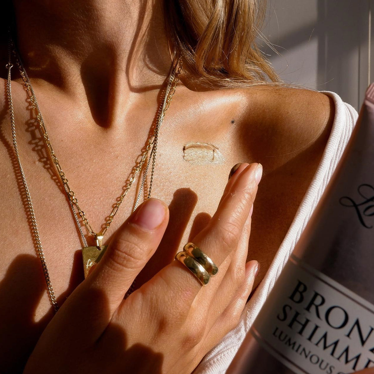 Eine Frau trägt Loving Tan Bronze Shimmer Luminous Cream auf ihr Schlüsselbein auf und trägt dazu Goldschmuck und ein weißes Oberteil.