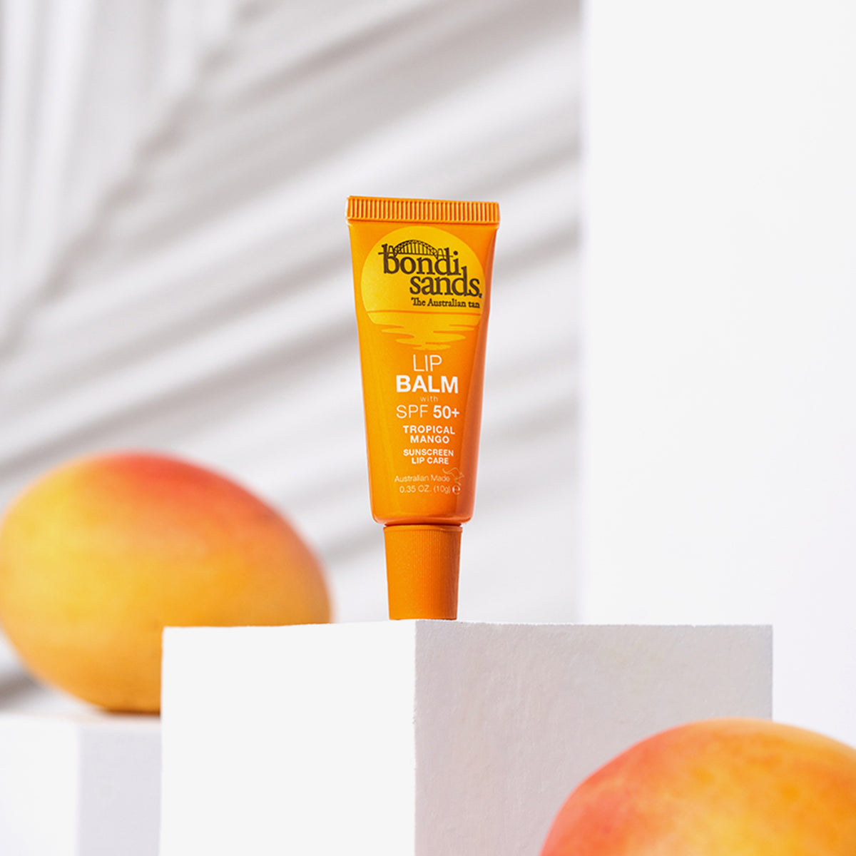 Eine Tube Bondi Sands Lip Balm SPF 50+ in Orange steht auf einem weißen Sockel, umgeben von Mangos