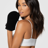 Eine Frau in einem weißen Oberteil hält den schwarzen Loving Tan Deluxe Self Tanning Applicator Mitt und blickt über ihre Schulter in die Kamera.