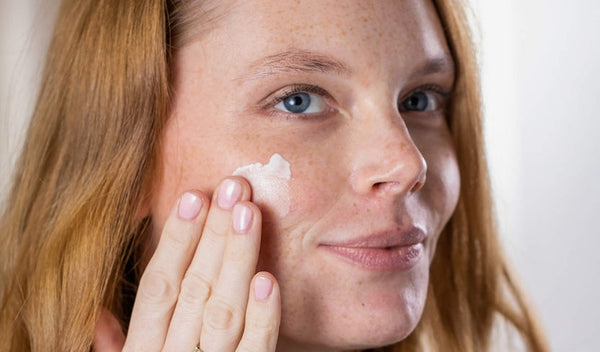 Frau mit Sommersprossen trägt Gesichtscreme auf ihre Wange auf und blickt leicht lächelnd in die Kamera.