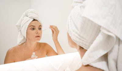 Frau mit Handtuchturban trägt vor dem Spiegel mit einer Pipette ein Pflegeprodukt auf ihr Gesicht auf – Pflegeroutine im Bad.