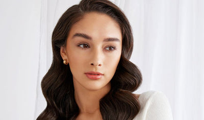 Eine Frau mit braunem welligem Haar und sanftem Make-up-Look vor weißen Vorhängen.