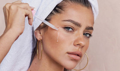Eine Frau mit Handtuch auf dem Kopf trägt Serum mit einer Pipette auf ihre Wange auf.