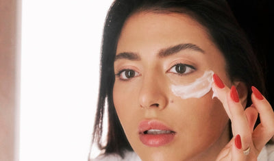 Frau mit dunklen Haaren beim Auftragen von Augencreme mit roten Fingernägeln.