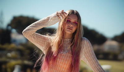 Frau mit langen Haaren und pinken Strähnen trägt Spitzenoberteil und auffälliges pinkes Augen-Make-up, steht mit geschlossenen Augen im Freien.