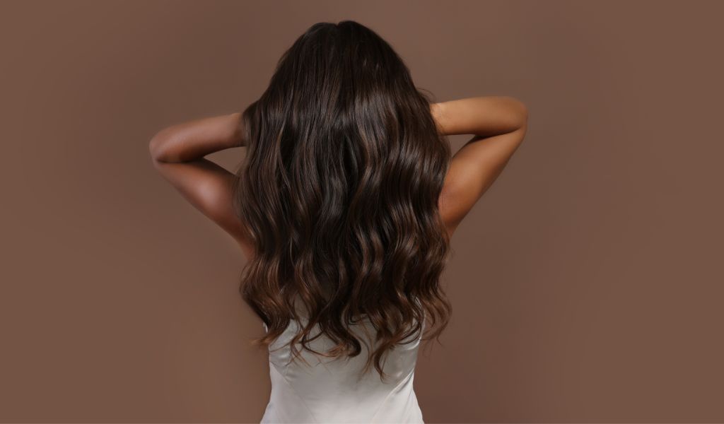 Frau mit langen, welligen, glänzenden braunen Haaren in weißem Top, von hinten vor braunem Hintergrund fotografiert.