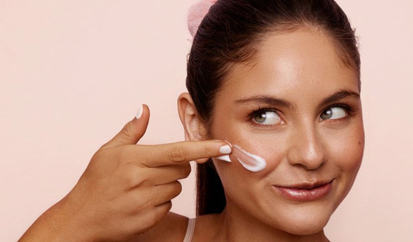 Frau trägt lächelnd Gesichtscreme auf ihre Wange auf und blickt zur Seite, vor rosafarbenem Hintergrund.