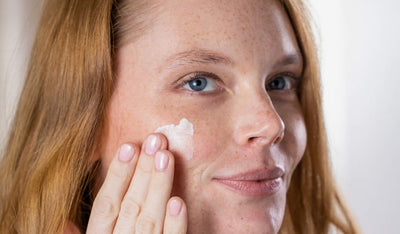 Eine Frau mit rotem Haar und blauen Augen lächelt, während sie Creme auf ihre Wange trägt.