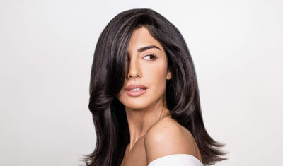 Frau mit glänzendem, voluminösem, dunkelbraunem Haar und Off-Shoulder-Oberteil blickt zur Seite vor hellem Hintergrund.