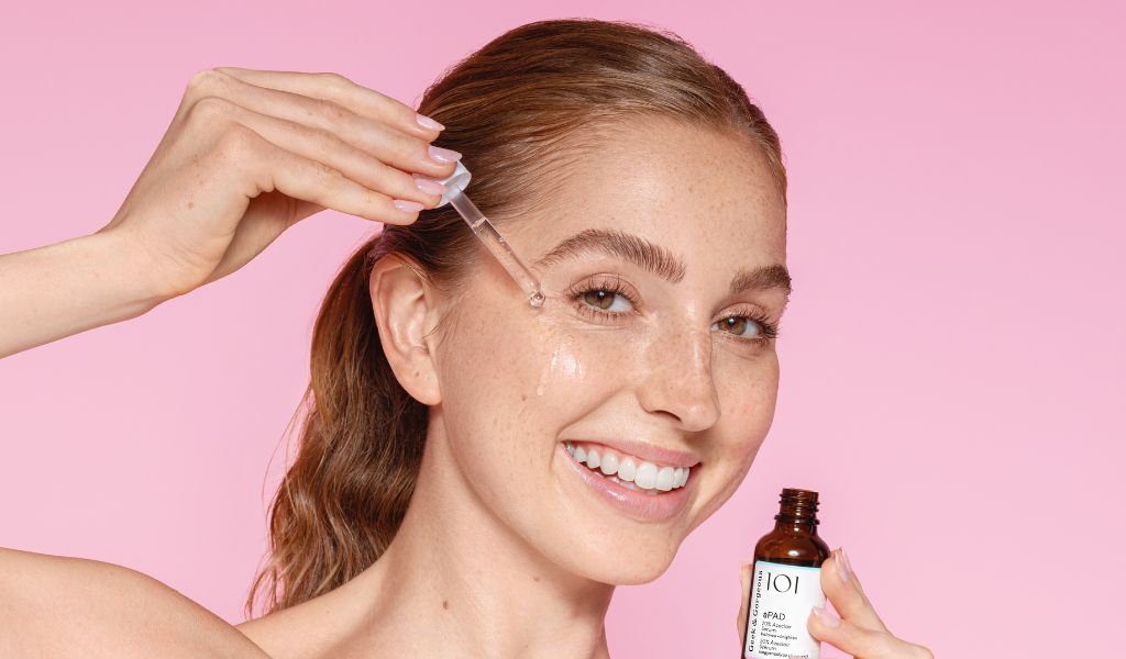 Lächelnde Frau trägt mit einer Pipette Gesichtsserum auf ihre Wange auf, vor rosa Hintergrund.