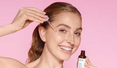 Lächelnde Frau trägt mit einer Pipette Gesichtsserum auf ihre Wange auf, vor rosa Hintergrund.