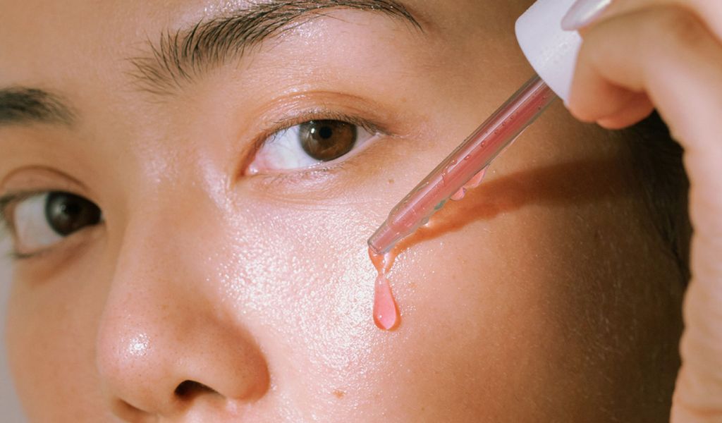 Nahaufnahme eines Gesichts, auf das eine rosafarbene Serum-Tropfen mit einer Pipette aufgetragen wird.