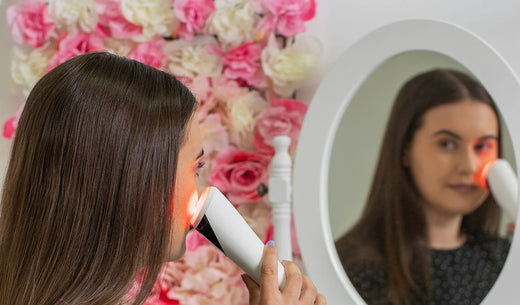 Frau verwendet ein rotes LED-Gerät zur Gesichtsbehandlung vor einem Spiegel, umgeben von rosa und weißen Blumen.