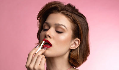 Frau mit kurzem braunem Haar trägt roten Lippenstift auf, Nahaufnahme vor rosa Hintergrund, Augen geschlossen, elegante Pose.