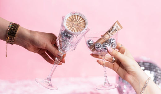 Zwei Hände halten Cocktailgläser gefüllt mit Mini-Diskokugeln und Make-up Produkten.