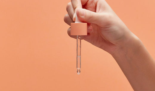 Hand hält eine Pipette mit durchsichtigem Serum vor orangem Hintergrund.