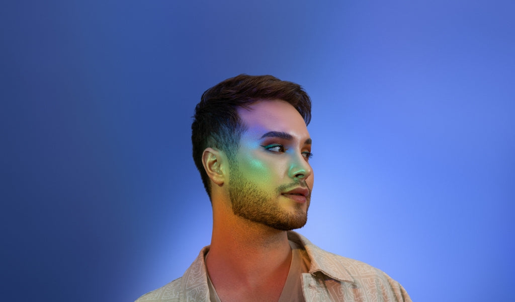  Ein Mann mit Bart und geschminkten Augen blickt nach rechts. Sein Gesicht wird von einem Regenbogenlicht beleuchtet.