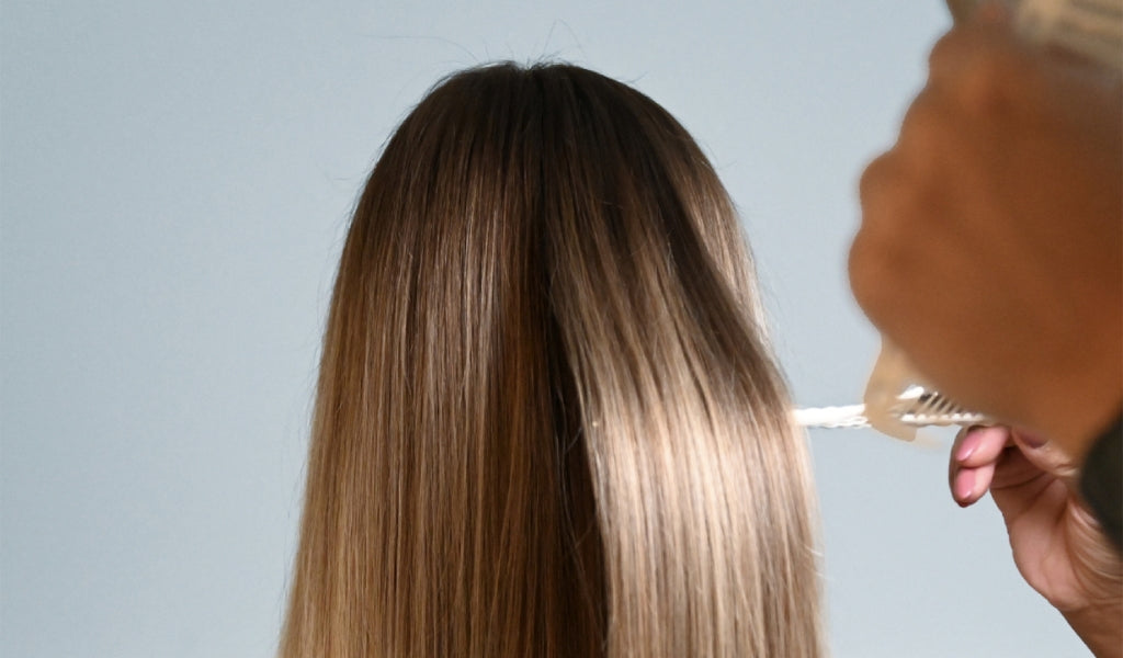 Glattes, braunes Haar von hinten, während ein Spray auf die Kopfhaut aufgetragen wird, vor neutralem Hintergrund.