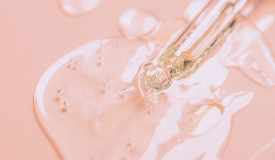 Nahaufnahme einer Pipette mit durchsichtigem Serum auf rosafarbenem Untergrund, mit glänzender Textur und kleinen Bläschen.