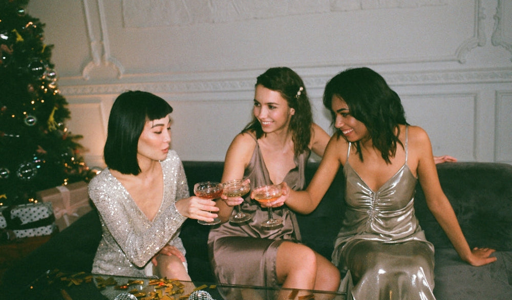 Drei elegant gekleidete Frauen stoßen mit Gläsern auf dem Sofa an, im Hintergrund steht ein geschmückter Weihnachtsbaum.