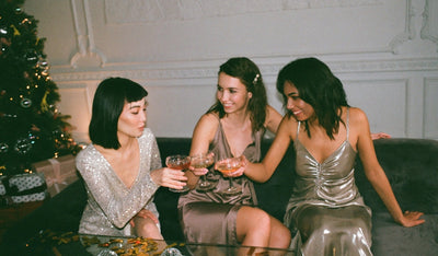 Drei elegant gekleidete Frauen stoßen mit Gläsern auf dem Sofa an, im Hintergrund steht ein geschmückter Weihnachtsbaum.