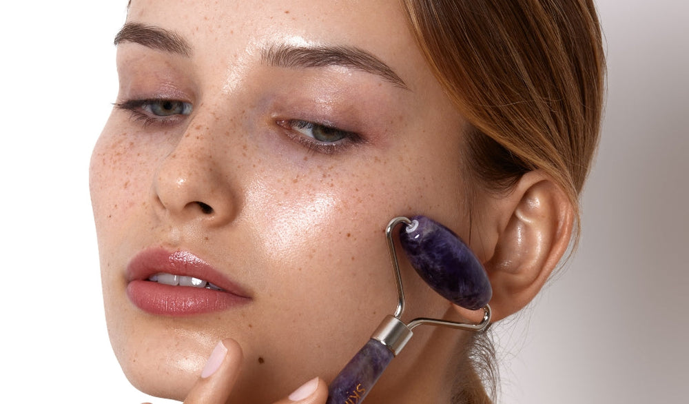Frau mit Sommersprossen nutzt einen violetten Gesichtsroller zur Hautpflege; ihre Haut glänzt frisch und gepflegt im Nahaufnahmebild.