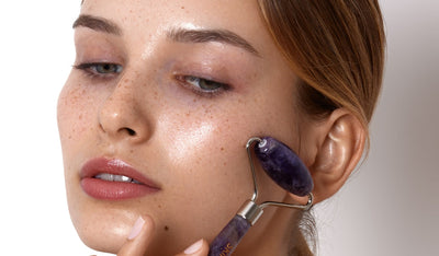 Frau mit Sommersprossen nutzt einen violetten Gesichtsroller zur Hautpflege; ihre Haut glänzt frisch und gepflegt im Nahaufnahmebild.