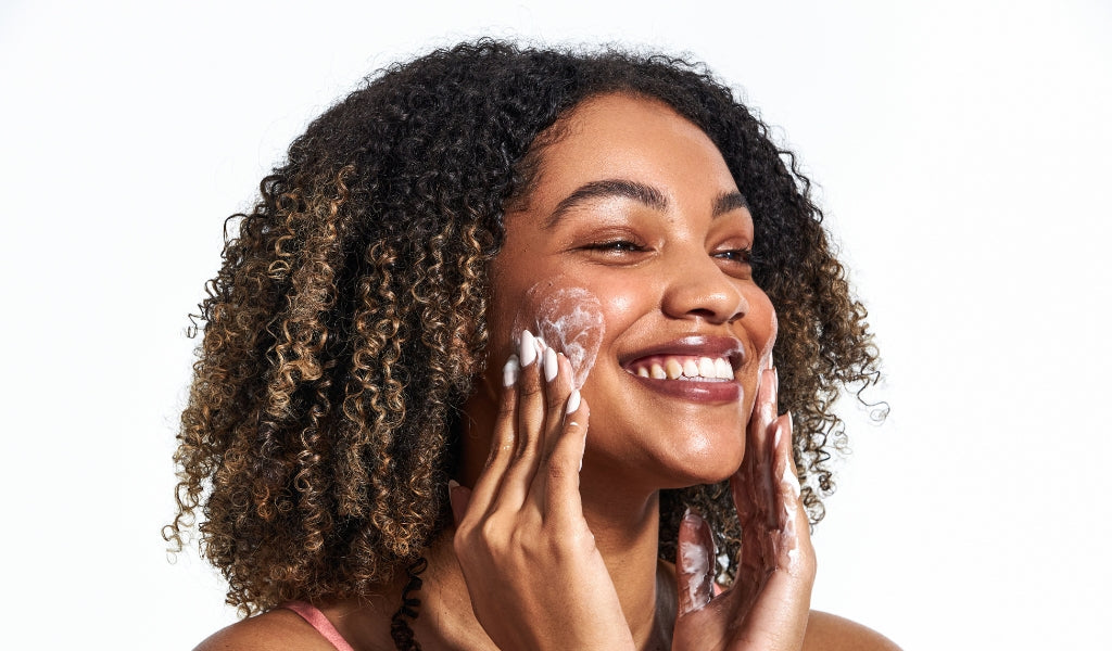Lächelnde Frau mit lockigem Haar trägt einen schäumenden Cleanser auf ihr Gesicht auf.