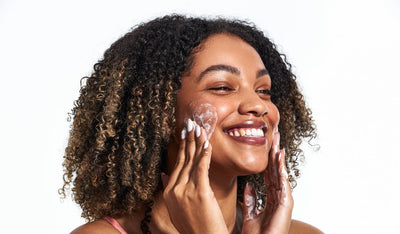 Lächelnde Frau mit lockigem Haar trägt einen schäumenden Cleanser auf ihr Gesicht auf.