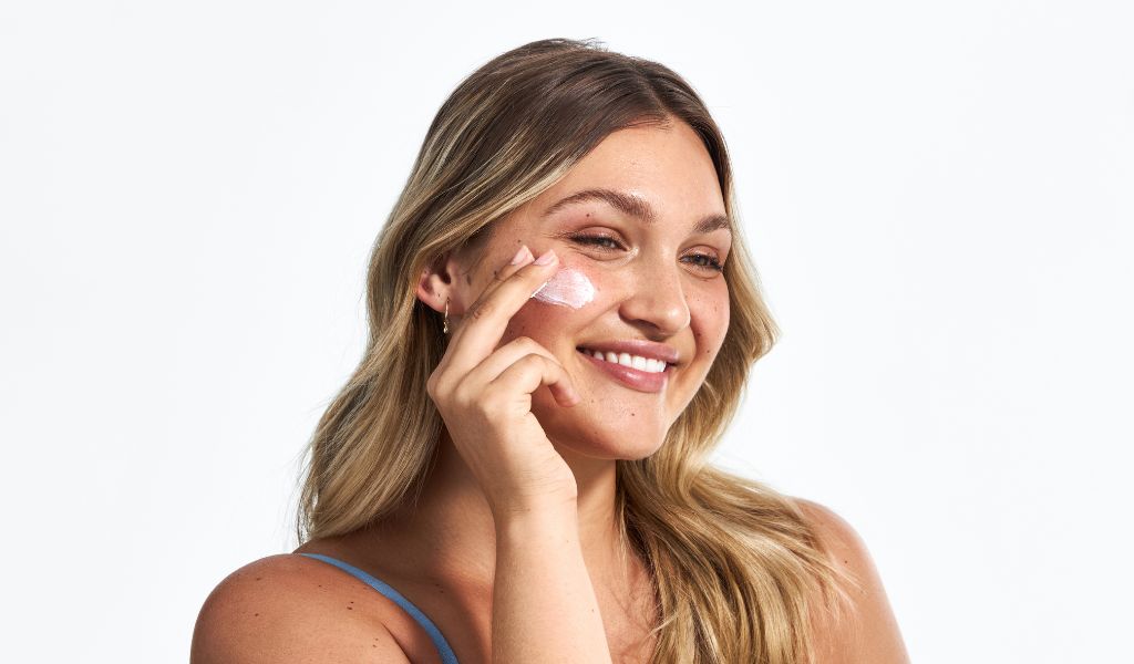 Lächelnde Frau trägt Gesichtscreme auf ihre Wange auf, vor hellem Hintergrund.