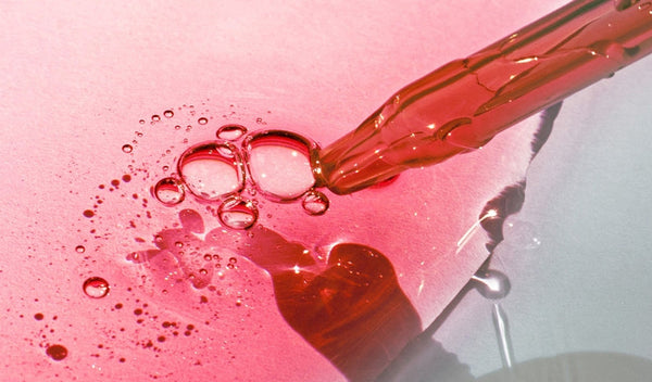 Rosa Serum tropft aus einer Pipette, umgeben von glänzenden Bläschen auf rosa Hintergrund.