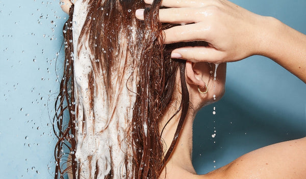 Person wäscht nasses Haar mit schäumendem Shampoo unter fließendem Wasser, Hand massiert die Kopfhaut.