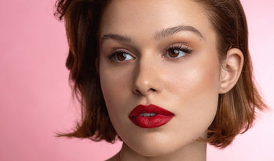 Frau mit kurzem braunem Haar, makellosem Teint und auffälligem roten Lippenstift vor rosa Hintergrund, elegant geschminkt.
