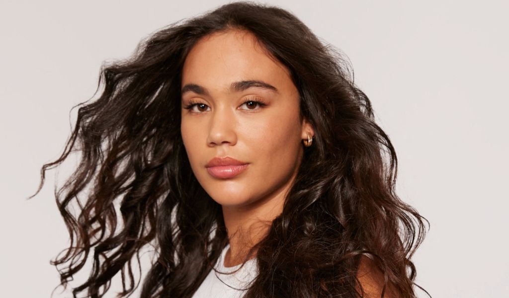 Frau mit langen, dunklen Locken und natürlichem Make-up blickt selbstbewusst in die Kamera, während ihr Haar dynamisch im Wind weht.