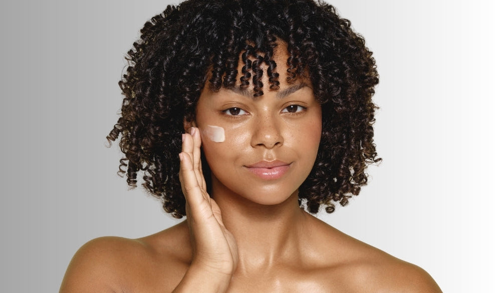 Frau mit lockigem Haar trägt sanft Gesichtscreme unter dem Auge auf, lächelt dezent, neutraler Hintergrund, natürliche Hautpflege.