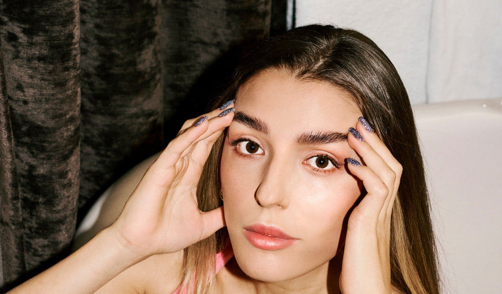 Frau mit glatten Haaren und natürlichem Make-up hält ihren Kopf mit beiden Händen und blickt direkt in die Kamera.