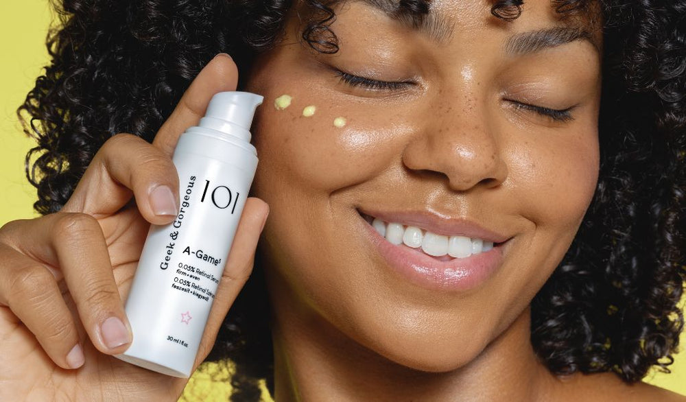 Frau mit Locken hält Retinol-Creme vor gelbem Hintergrund und trägt Produkt lächelnd auf ihre Wange auf.