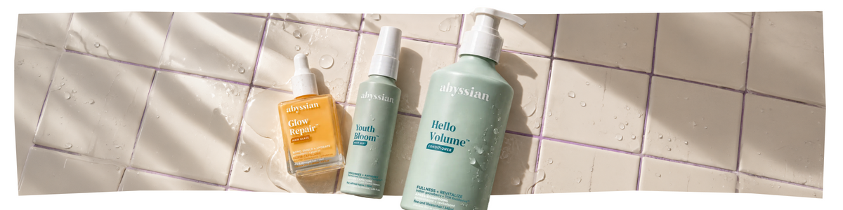 Drei Haarpflegeprodukte von Abysian – „Glow Ritual“, „Youth Bloom“ und „Hello Volume“ – stehen an den nassen, beige gefliesten Duschwänden, beleuchtet vom Sonnenlicht und übersät mit Wassertropfen.
