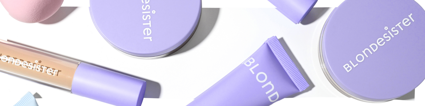 Blondesister-Make-up-Produkte, arrangiert auf weißem Hintergrund, darunter eine Puderdose, eine Tube, ein Schwämmchen und ein Concealer, alle in einheitlicher lavendelfarbener Verpackung mit weißer Beschriftung.