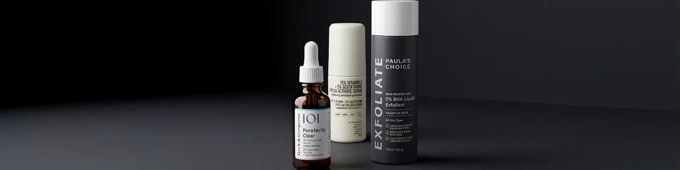 Drei Hautpflegeflaschen, darunter eine Tropfflasche, angeordnet auf einer dunklen Fläche mit dunklem Hintergrund.