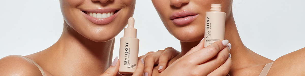 Zwei lächelnde Frauen mit Bali Body Serum-Flaschen, beide mit strahlendem, natürlichem Make-up.
